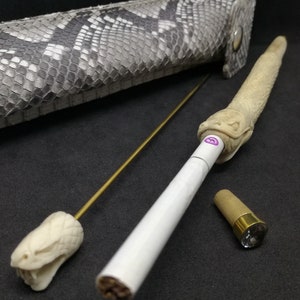 May include: A white cigarette with a snake-shaped wooden mouthpiece, a gold metal rod with a snake-shaped wooden tip, and a brown wooden cap with a clear crystal on top. The items are on a black surface.
