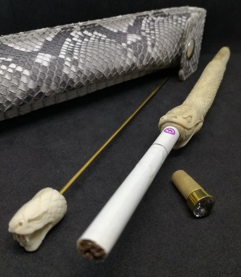 May include: A white cigarette with a snake-shaped wooden mouthpiece, a gold metal rod with a snake-shaped wooden tip, and a brown wooden cap with a clear crystal on top. The items are on a black surface.