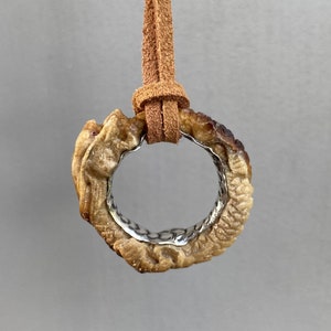 Puede incluir: Un collar de cordón de cuero marrón con un colgante de hueso tallado. El colgante tiene forma de círculo y presenta un diseño detallado.