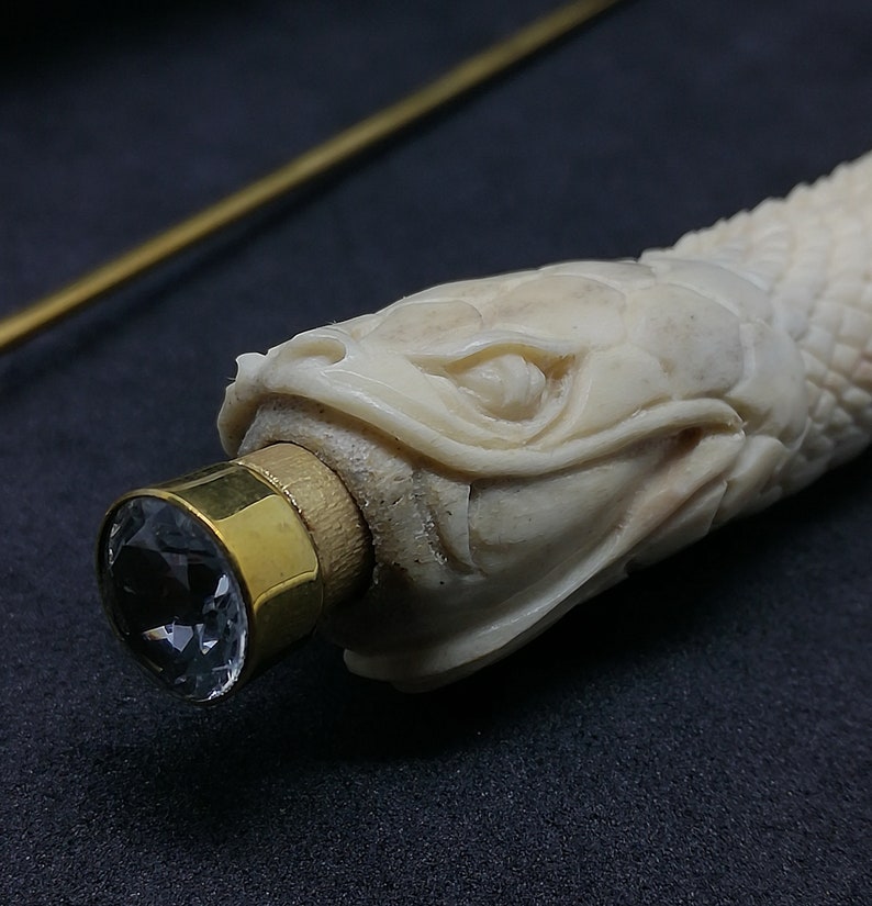 May include: A white bone carving of a fish head with a brass cap and a clear crystal on the end.