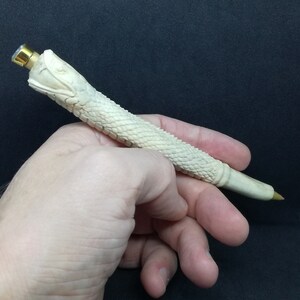 May include: A white, carved pen shaped like a snake with a gold-coloured top. The pen is held in a person's hand.