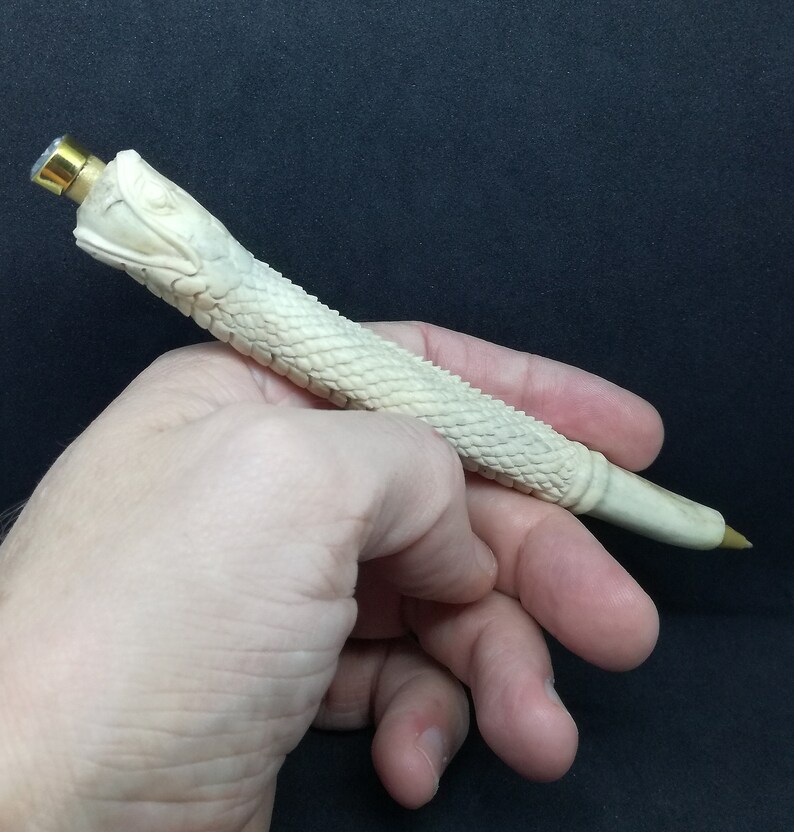 May include: A white, carved pen shaped like a snake with a gold-coloured top. The pen is held in a person's hand.