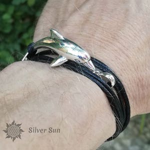 May include: A black leather bracelet with a silver dolphin charm. The bracelet is layered and has a silver clasp. The text "Silver Sun" is visible on the wrist.