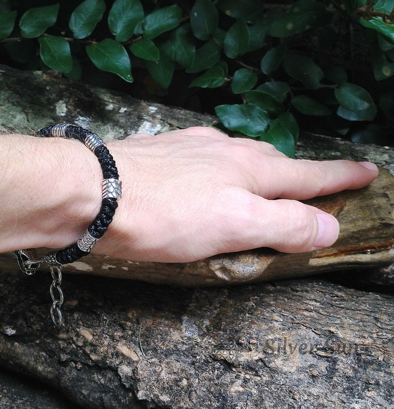 Puede incluir: Un brazalete de cuero negro con detalles plateados. El brazalete tiene un dise&ntilde;o trenzado y un cierre de broche.