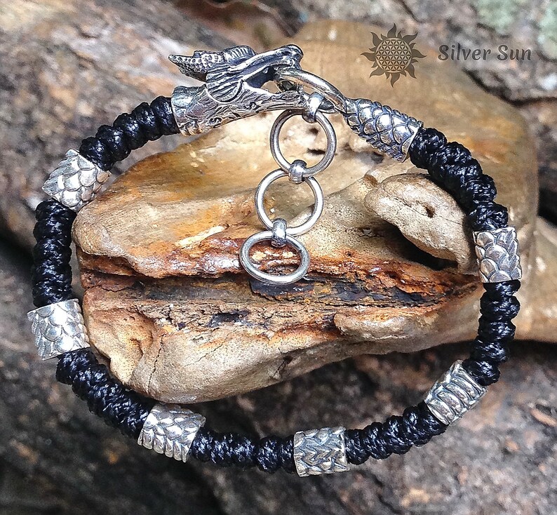 Puede incluir: Pulsera trenzada negra con cierre de cabeza de drag&oacute;n plateado y cuentas plateadas. Las cuentas tienen un patr&oacute;n de escamas. La pulsera est&aacute; sobre una superficie de madera. Silver Sun.
