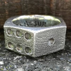 Op de afbeelding: Zilverkleurige ring in de vorm van een dobbelsteen, met getextureerde oppervlakken en cirkelvormige inkepingen die de getallen één tot en met zes voorstellen. De ring heeft een vierkante vorm en een matte afwerking, geschikt voor dagelijks gebruik.
