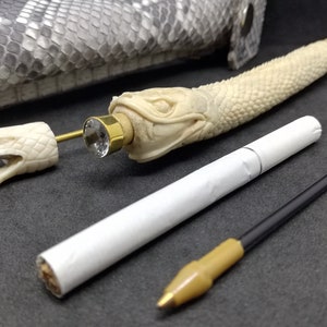 May include: A white snake-shaped pipe with a gold-coloured mouthpiece and a clear crystal embellishment. The pipe is lying on a grey textured surface with a white cigarette and a black and brown cord.