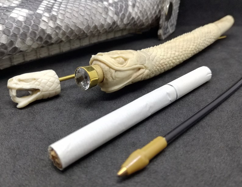 May include: A white snake-shaped pipe with a gold-coloured mouthpiece and a clear crystal embellishment. The pipe is lying on a grey textured surface with a white cigarette and a black and brown cord.