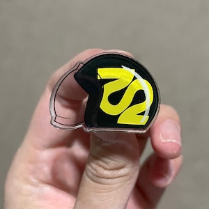 Pode incluir: Um alfinete de plástico transparente com um design preto e amarelo de um capacete com as letras "RS" nele.