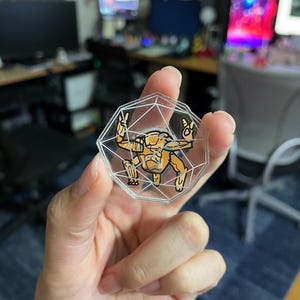 May include: A clear, multi-faceted acrylic token shaped like a twenty-sided die. Inside the transparent geometric frame is a printed illustration of an orange, robotic-looking creature with multiple limbs, making a peace sign with its hands.