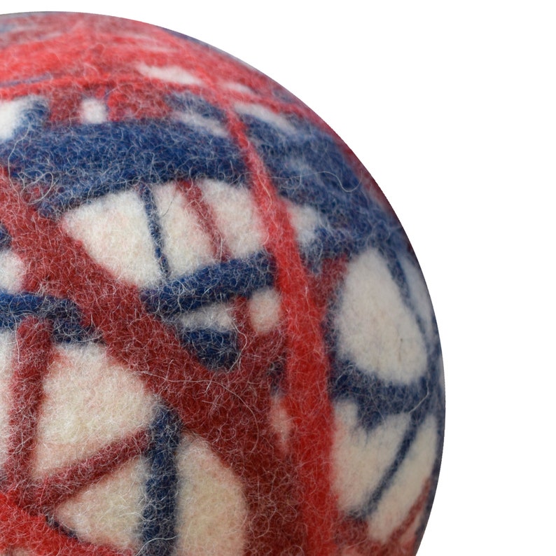 Puede incluir: Una bola de fieltro blanca, azul y roja con un patr&oacute;n geom&eacute;trico. La bola est&aacute; hecha de lana y tiene una textura suave y peluda.