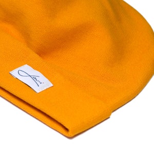 Puede incluir: Primer plano de un gorro de color amarillo mostaza. El gorro tiene una textura suave y una peque&ntilde;a etiqueta blanca con texto cursivo azul. El gorro est&aacute; hecho de tejido de punto y es de color s&oacute;lido.