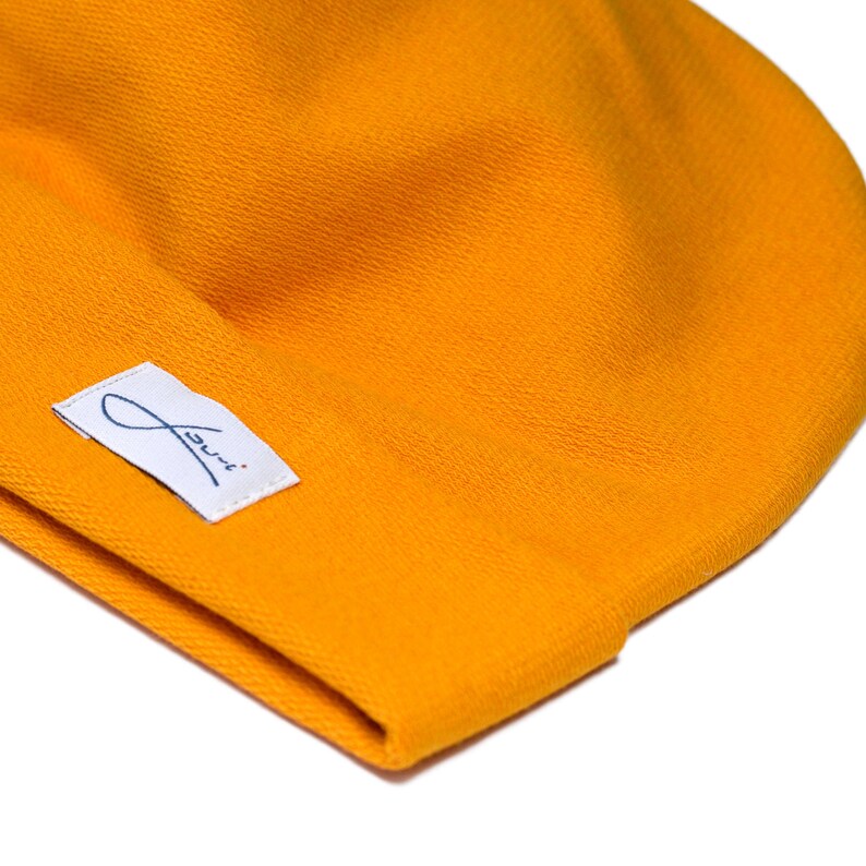 Puede incluir: Primer plano de un gorro de color amarillo mostaza. El gorro tiene una textura suave y una peque&ntilde;a etiqueta blanca con texto cursivo azul. El gorro est&aacute; hecho de tejido de punto y es de color s&oacute;lido.