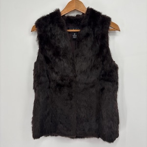 May include: A dark brown fur vest with a V-neck design. The vest is hanging on a wooden hanger against a white background. The fur appears soft and plush, with a natural texture.