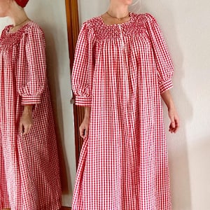 May include: A floor-length red and white gingham dress featuring a square neckline with smocked detailing, long puff sleeves with buttoned cuffs, and a loose, flowing silhouette. A matching red bandana is worn as a headpiece with the dress.