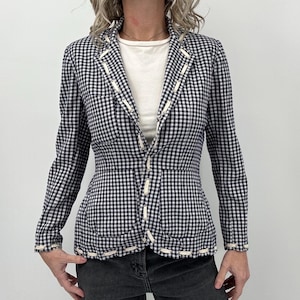 May include: A black and white gingham blazer with white trim and a fitted silhouette. The blazer has long sleeves and a button-up front. The trim is stitched along the edges of the blazer, pockets, and lapels. The blazer is paired with a white top and dark gray jeans.