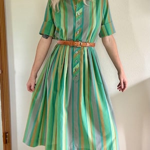 May include: A midi-length shirt dress featuring vertical stripes in shades of green, grey, and tan. The dress has short sleeves with folded cuffs, a collared neckline, a button-down front placket, and a thin brown leather belt with a gold buckle.