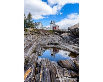 May include: A white lighthouse with a red roof stands on a rocky shore. The lighthouse is reflected in a small pool of water.