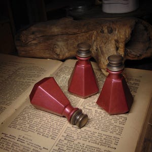 May include: Three red, faceted glass bottles with brass-colored tops rest on an open book. One bottle is lying on its side. The bottles have a vintage aesthetic.