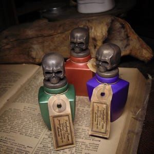 May include: Three decorative bottles with skull-shaped stoppers. The bottles are in green, red, and purple, each with a tag that reads "Paralysing Poison". They rest on an open book with aged pages, evoking a vintage apothecary theme.