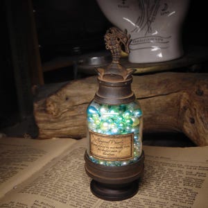 May include: A clear glass bottle filled with iridescent blue and green pearls, featuring a dark brown metal base and a matching lid topped with a seahorse figurine. A rectangular label on the front reads Abyssal Pearls hand picked by mermaids from the deepest parts of the ocean.