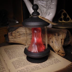 May include: A glass jar with a black base and lid, holding a red, shapeless form. The jar rests on an open book with aged pages. A white ceramic hand with palmistry markings is in the background, with a piece of driftwood.