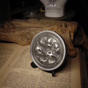 May include: A silver-colored, circular object with an uneven, textured surface, possibly a lid or decorative piece. It rests on a stand, placed on an open book with German text. A white ceramic object with text is in the background.