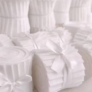 May include: Several white fabric bundles, some rolled and others stacked, secured with white satin ribbons. The fabric is folded and layered, giving a textured look. The image suggests a craft or sewing supply, possibly for quilting.