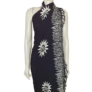 May include: A dark navy blue sarong featuring a white edelweiss floral pattern and a vertical side panel with a white wavy striped design. The fabric edge is finished with a fringe detail, creating a textured look along the side of the garment.