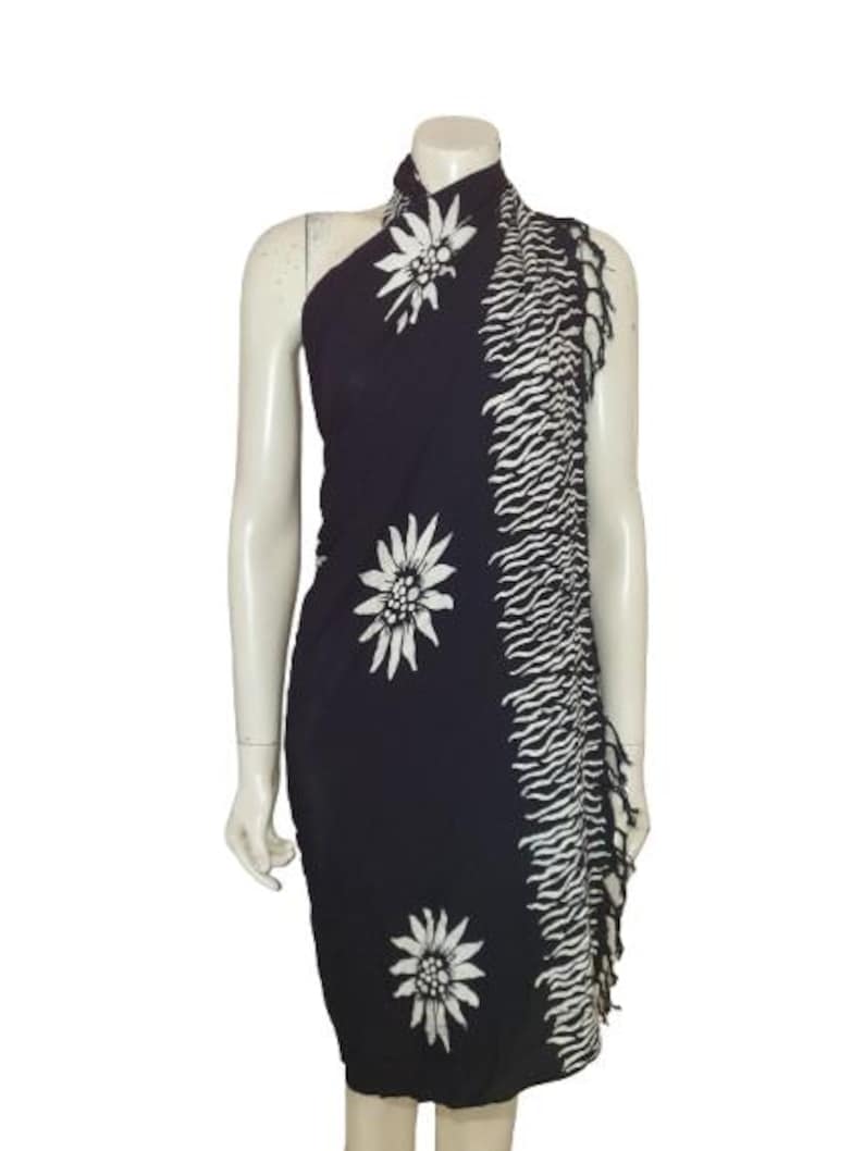 May include: A dark navy blue sarong featuring a white edelweiss floral pattern and a vertical side panel with a white wavy striped design. The fabric edge is finished with a fringe detail, creating a textured look along the side of the garment.