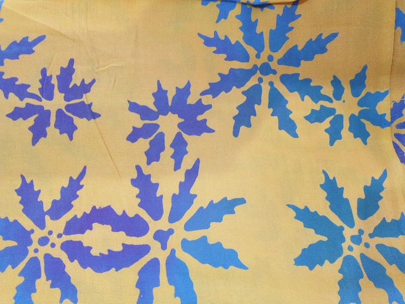 May include: A mustard yellow fabric featuring a repeating pattern of stylized blue floral shapes. The flowers have jagged, leaf-like petals radiating from a central point, creating a bold contrast against the warm, muted yellow background.