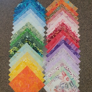 May include: A stack of 20 colourful fabric squares with various patterns, including floral, geometric, and abstract designs. The squares are arranged in a rainbow pattern, with blue, green, yellow, orange, and red squares on the left and purple, pink, and brown squares on the right.