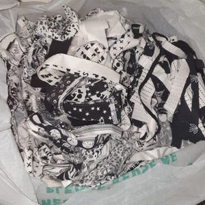 May include: A transparent plastic bag overflowing with black and white fabric remnants. The fabrics display diverse patterns, such as stars, geometric designs, and text. The bag is partly open, showing the contents.