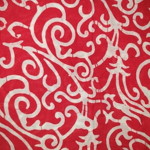 May include: A vibrant red fabric featuring a continuous, repeating off-white pattern of intricate, flowing, and interconnected scrollwork designs. The surface displays a subtle, cracked texture throughout the white lines, characteristic of batik.