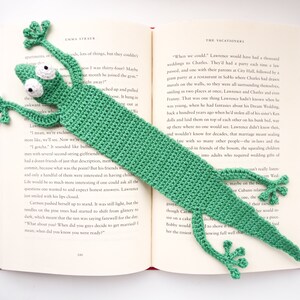 Pu&ograve; includere: Un segnalibro a forma di geco verde all'uncinetto con un viso dettagliato e una lunga coda. Il segnalibro &egrave; posizionato su un libro aperto con testo sui matrimoni.