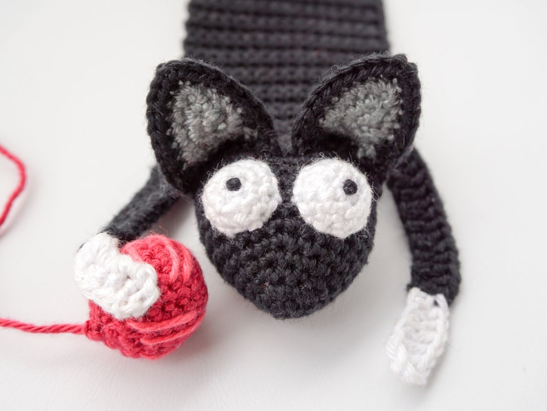 May include: A black crocheted cat bookmark with white yarn accents and large white eyes. The cat is holding a red yarn ball.