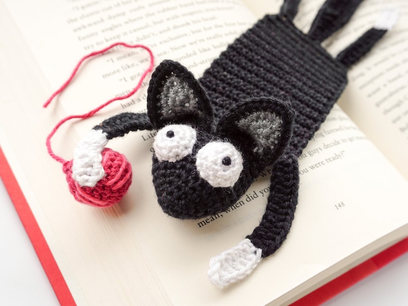 May include: A black crocheted cat bookmark with white yarn accents and large white eyes. The bookmark is holding a small red yarn ball.