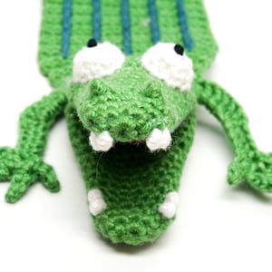 Puede incluir: Marioneta de cocodrilo verde de crochet con dientes blancos y grandes ojos blancos. La marioneta tiene la boca muy abierta.
