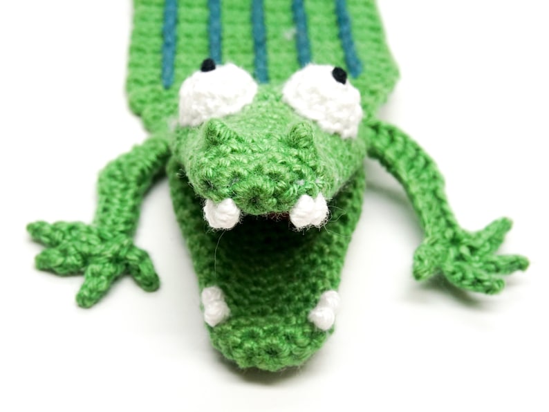 Puede incluir: Marioneta de cocodrilo verde de crochet con dientes blancos y grandes ojos blancos. La marioneta tiene la boca muy abierta.