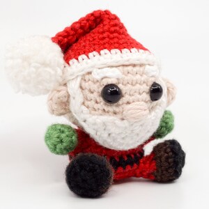 Puede incluir: Un adorno de Pap&aacute; Noel de crochet con un gorro rojo, barba blanca y ojos negros. El adorno est&aacute; sentado sobre una superficie blanca.