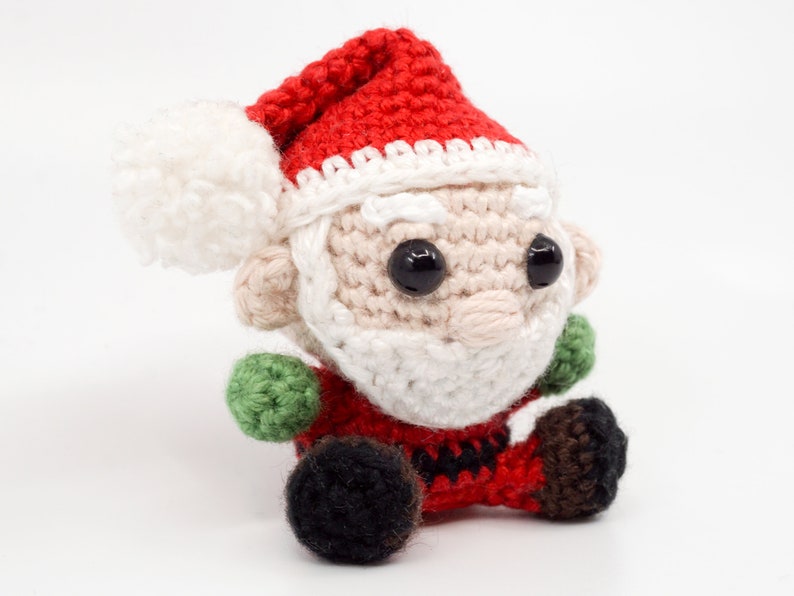 Puede incluir: Un adorno de Pap&aacute; Noel de crochet con un gorro rojo, barba blanca y ojos negros. El adorno est&aacute; sentado sobre una superficie blanca.