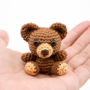 May include: A small brown crocheted teddy bear with black eyes and a brown nose. The bear is sitting in the palm of a hand.