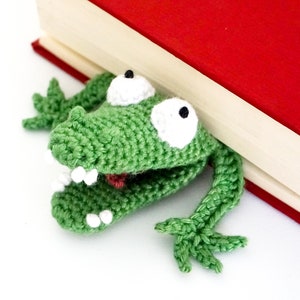 Puede incluir: Marcap&aacute;ginas de rana de ganchillo verde con ojos y dientes blancos, asom&aacute;ndose por detr&aacute;s de un libro rojo.