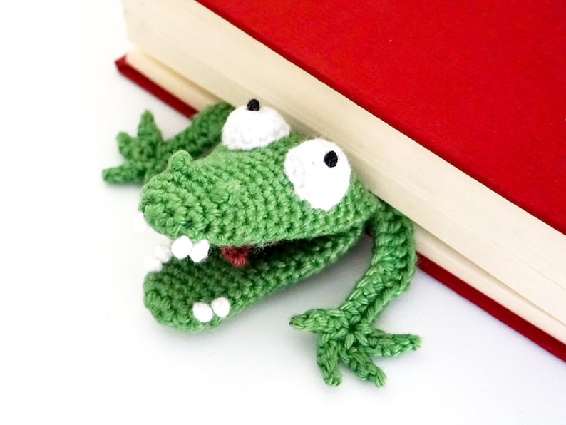 Puede incluir: Marcap&aacute;ginas de rana de ganchillo verde con ojos y dientes blancos, asom&aacute;ndose por detr&aacute;s de un libro rojo.
