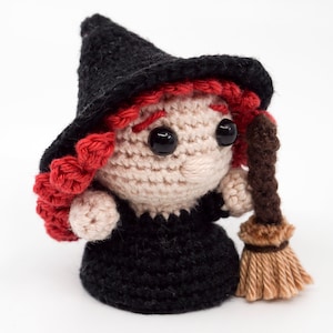 May include: A crocheted witch doll with red hair, a black hat, and a brown broom. The doll is wearing a black dress.