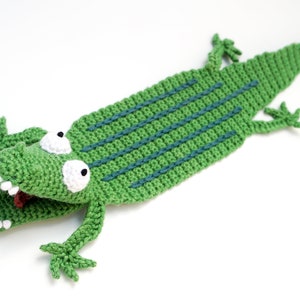 Puede incluir: Marcap&aacute;ginas de cocodrilo verde tejido a crochet con ojos blancos y lengua roja. El cocodrilo tiene una espalda texturizada con rayas azules.