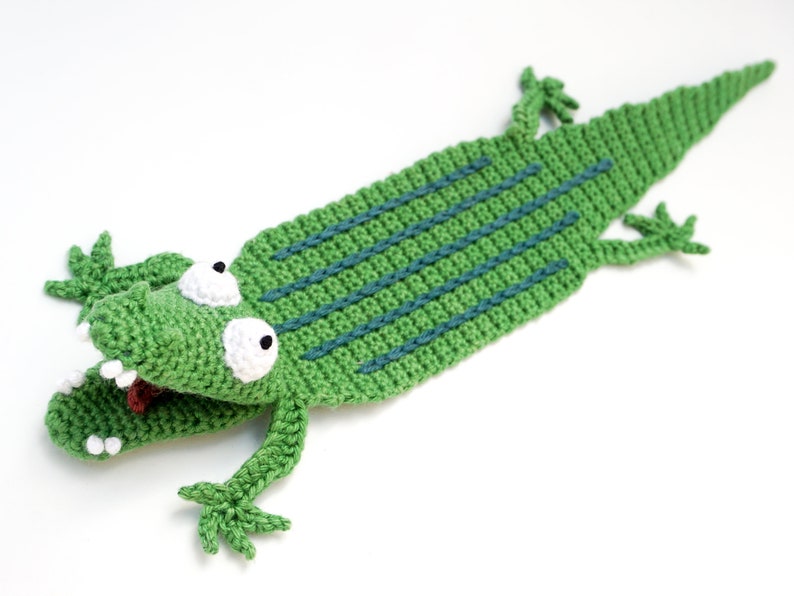 Puede incluir: Marcap&aacute;ginas de cocodrilo verde tejido a crochet con ojos blancos y lengua roja. El cocodrilo tiene una espalda texturizada con rayas azules.