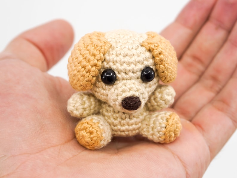 May include: A small crocheted dog toy with brown eyes, a brown nose, and beige and brown fur.