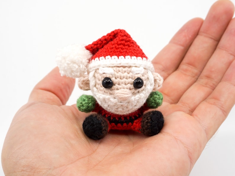 Puede incluir: Un peque&ntilde;o mu&ntilde;eco de Pap&aacute; Noel de crochet que lleva un gorro rojo con ribete blanco y una barba blanca. El mu&ntilde;eco est&aacute; sentado con las piernas cruzadas y lleva guantes verdes.