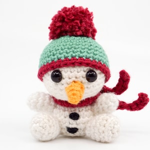 Puede incluir: Un muñeco de nieve de crochet que lleva un gorro verde y rojo con un pompón rojo y una bufanda roja. El muñeco de nieve tiene ojos negros, una nariz naranja y botones negros.